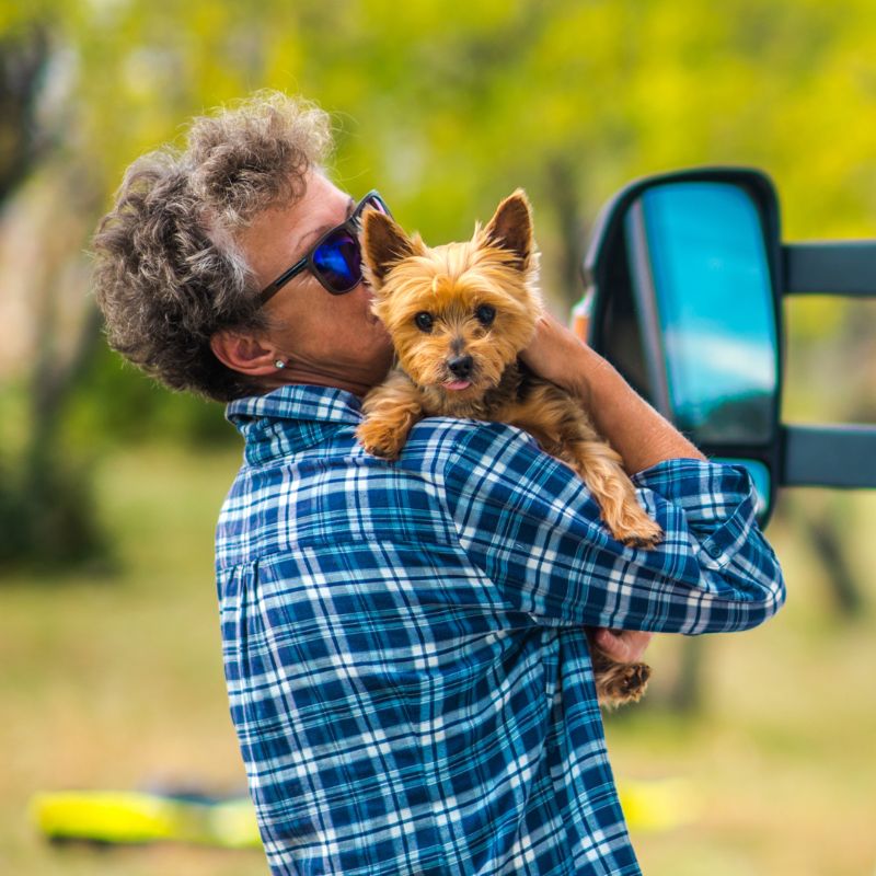 retired woman wearing a blue plaid shirt holds her Yorkie dog while standing next to a van